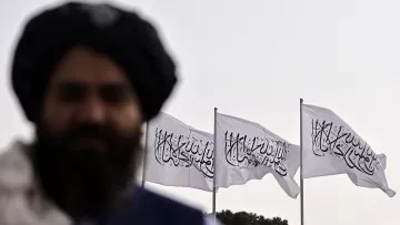 L'immagine mostra un uomo non completamente a fuoco in primo piano, con una folta barba e un turbante. Sullo sfondo, ci sono tre bandiere bianche che sventolano, ognuna recante un simbolo o una scritta. Le bandiere evocano un contesto culturale e politico specifico, tipico di alcune regioni. L'atmosfera è suggestiva e mette in risalto il contrasto tra il soggetto in primo piano e lo sfondo.