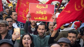 تظهر الصورة مجموعة من الأشخاص في مسيرة احتجاجية. يحمل أحد المتظاهرين لافتة مكتوب عليها "الحرية للولد الشعب"، بينما يبدو أن الحشود من حوله ترفع أعلامًا حمراء. التعبيرات في وجوه المتظاهرين تتراوح بين الحماس والعزيمة، مما يدل على روح التضامن والمشاركة في المطالبة بالحقوق. الجو العام يظهر فيه نشاط وحيوية، مما يشير إلى أهمية الرسالة التي يسعون لنقلها.