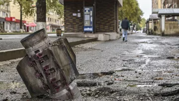 L'image montre un paysage urbain marqué par la destruction. Au premier plan, on voit un obus ou un engin explosif détérioré, partiellement enfoui dans le sol. Le bitume est endommagé autour de l'obus, laissant des fissures et des débris. En arrière-plan, un homme marche sur le trottoir, près d'un bâtiment qui semble intact. L'atmosphère est sombre et témoigne des conséquences d'un conflit ou d'un événement violent. Les arbres sur le bord de la route ajoutent une touche de verdure, mais le contexte reste très préoccupant.