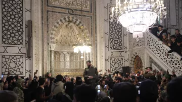 در تصویر، گروهی از افراد در یک مکان مذهبی یا تاریخی تجمع کرده‌اند. در وسط تصویر، فردی با لباس نظامی در حال سخنرانی یا بیان مطلبی به جمعیت است. نفرات در اطراف او با تلفن‌های همراه خود مشغول فیلم‌برداری یا عکاسی هستند. محیط اطراف آن‌ها تزئینات زیبا و نورپردازی چشمگیری دارد، همچنین یک چراغ آویز بزرگ در بالای سر گروه دیده می‌شود که به فضا زیبایی می‌بخشد. دیوارها با نقوش و طراحی‌های پیچیده تزئین شده‌اند و جو عمومی تصویر نشان‌دهنده شور و شوق جمعیت است.