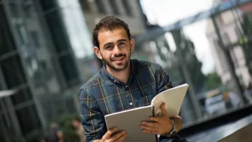 في الصورة، يظهر شاب يجلس في مكان يبدو حديثًا وعصريًا. يرتدي قميصًا مزخرفًا مربعات وبنطال جينز. يحمل دفترًا مفتوحًا في يده، ويبدو مبتسمًا وسعيدًا، مما يوحي بأنه في لحظة من التركيز أو الدراسة. خلفه يمكن رؤية بنايات زجاجية وأشجار، ما يعطي انطباعًا عن بيئة حضرية نهارية.