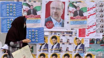 در این تصویر، یک زن که با چادر مشکی پوشیده است، در حال عبور از کنار دیواری است که پر از پوسترهای انتخاباتی و تبلیغاتی است. پوسترها شامل تصاویری از نامزدهای مختلف و شعارهای انتخاباتی هستند. چهره‌ای از یک فرد (احتمالاً نامزد) در مرکز تصویر بسیار مشهود است، در حالی که دیگر پوسترها در اطراف آن به چشم می‌خورند. دیوار پر از رنگ و متن است که به فضای انتخاباتی اشاره می‌کند. زن در حال نگاه کردن به این پوسترها به نظر می‌رسد.
