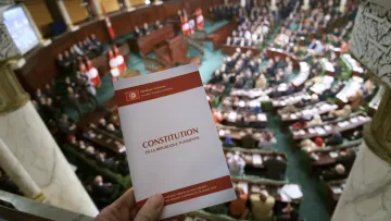 The image shows a parliamentary session in Tunisia, with a focus on a hand holding a book titled "Constitution de la République Tunisienne" (Constitution of the Tunisian Republic). In the background, a large assembly of representatives is seated in a formal, legislative setting, likely discussing or voting on matters related to the constitution or other important legislative issues. The architecture of the chamber features ornate details, and national flags are visible, emphasizing the official nature of the event.