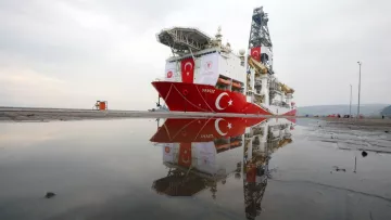 L'image montre un navire de forage maritime, nommé "Yavuz", qui est amarré au port. Le bateau arbore des couleurs dominantes de rouge et blanc, représentant le drapeau turc. On peut également voir des équipements de forage sur le pont du navire. Dans la partie inférieure de l'image, il y a des réflexions du navire dans une flaque d'eau sur le sol. Le ciel en arrière-plan est nuageux, créant une atmosphère calme.