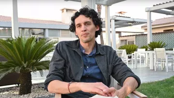 L'image montre un homme assis sur une chaise en bois dans un environnement extérieur. Il a des cheveux bouclés et porte une chemise sombre sur un t-shirt. Il est assis dans un cadre paisible, avec des plantes vertes autour de lui et une terrasse visible en arrière-plan. L'ambiance semble détendue et agréable.