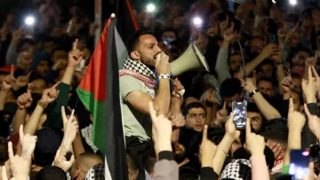 L'image montre un grand rassemblement de personnes, probablement lors d'une manifestation. Au premier plan, un homme utilisant un mégaphone pour parler à la foule. Il est entouré de nombreux participants qui lèvent les bras et brandissent des drapeaux, notamment des drapeaux palestiniens. L'atmosphère semble électrisante et engagée, avec des gens qui filment l'événement avec leurs téléphones. La scène dénote une forte mobilisation et un sentiment collectif.