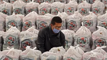 L'image montre un homme en train de travailler au milieu d'un grand espace rempli de sacs en plastique. Chaque sac est soigneusement empilé, arborant un motif de rose et des inscriptions en arabe. L'homme porte un masque et semble concentré sur sa tâche, probablement en train de trier ou de préparer quelque chose à l'intérieur des sacs. L'environnement est bien organisé, et la scène évoque un sentiment d'efficacité dans un contexte d'entraide ou de distribution.