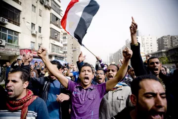 L'image montre une foule de manifestants rassemblés dans une rue urbaine. Certains d'entre eux lèvent les bras en l'air et expriment des émotions vives, comme la colère ou l'enthousiasme. Un homme au premier plan tient un drapeau égyptien bien en vue, symbole de la manifestation. Les bâtiments environnants montrent un décor urbain d'une grande ville, probablement Le Caire. L'ambiance générale suggère un moment de protestation ou de revendication collective.