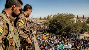 The image depicts a large crowd participating in a demonstration or march. In the foreground, two armed soldiers in military uniforms are positioned on a higher vantage point, observing the event. Below them, a diverse group of people holds various flags and banners, indicating that the gathering is likely a political or social protest. The scene is set in an outdoor urban environment, with trees and buildings visible in the background. The atmosphere appears to be one of engagement and collective action.