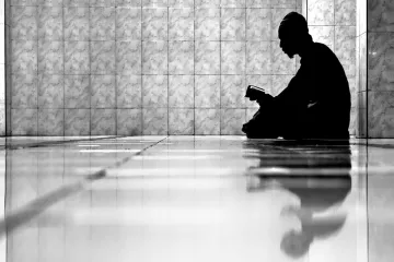 L'image montre un individu assis sur le sol, plongé dans la lecture d'un livre. La composition est en noir et blanc, créant un contraste fort entre l'ombre de la personne et l'éclat des carreaux brillants au sol. L'ambiance est calme et contemplative, et le reflet de la silhouette sur le sol ajoute une dimension introspective à la scène. Les murs en arrière-plan sont recouverts de carrelage, ce qui renforce l'aspect minimaliste de l'image.
