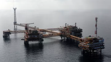 L'image montre une plateforme pétrolière en mer. On peut voir plusieurs structures métalliques assemblées, avec des passerelles reliant les différentes sections. Il y a des installations pour l'extraction du pétrole, ainsi que des antennes et des équipements de communication. La mer environnante est calme, et le ciel est partiellement nuageux. Cette scène évoque l'industrie pétrolière et offshore, avec une infrastructure conçue pour exploiter les ressources sous-marines.