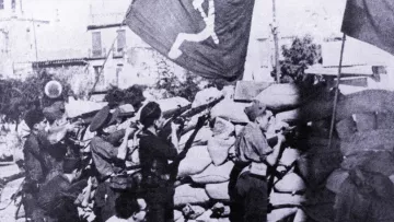 Cette image en noir et blanc montre un groupe de soldats ou de miliciens en position de tir, probablement lors d'un conflit. Ils se trouvent derrière un mur de sacs de sable, tout en tenant des fusils. En arrière-plan, des drapeaux avec un symbole communiste flottent, ce qui suggère un contexte politique et historique marqué par des idéologies de gauche. L'ambiance de la scène semble intense et sérieuse, indiquant un moment de bataille ou de lutte.
