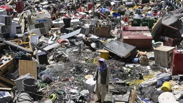 The image shows a cluttered scene filled with a massive amount of discarded items, including various types of debris, appliances, and objects that appear to be from different categories. Amidst this chaotic environment, a person is seen walking through the debris, navigating the piles of junk. The area looks like a dump or recycling site, emphasizing the vast accumulation of waste and the struggle to find salvageable materials within.