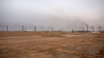 این تصویر یک محیط صنعتی را نشان می‌دهد که در آن دود از دودکش‌های بلند بیرون می‌آید. آسمان به طور کلی ابری و خاکستری به نظر می‌رسد و هوای منطقه آلوده است. زمین خاکی و خشک به نظر می‌رسد و هیچ گونه پوشش گیاهی قابل مشاهده نیست. این صحنه می‌تواند نشان‌گر فعالیت‌های صنعتی و مشکلات زیست‌محیطی باشد.