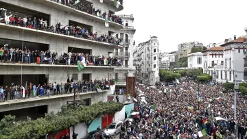 L'image montre une grande foule rassemblée devant un bâtiment, manifestant dans une ville. De nombreuses personnes sont visibles sur les balcons du bâtiment, brandissant des drapeaux et exprimant leur soutien à la manifestation. La rue est remplie de manifestants, et l'atmosphère semble vibrante et dynamique, avec un mélange de drapeaux et de pancartes. Le paysage urbain environnant, composé de divers bâtiments, ajoute à l'environnement de la scène.
