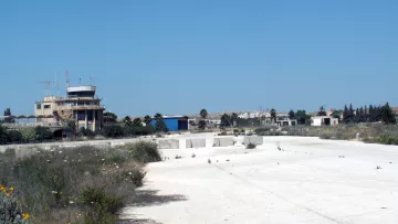 La imagen muestra un paisaje de un área desierta, posiblemente un antiguo aeropuerto o base aérea. En el fondo se observa una torre de control o edificio administrativo, rodeado de vegetación y estructuras en desuso. El suelo es de color claro y parece estar cubierto de hierbas y flores silvestres, lo que sugiere abandono. El cielo es de un azul despejado, lo que le da un contraste al paisaje.