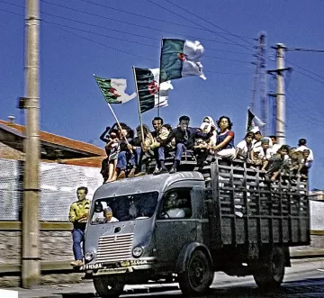 L'image montre un camion dont le plateau est rempli de personnes célébrant avec des drapeaux algériens. Les gens semblent joyeux et enthousiastes, tenant les drapeaux en l'air. En arrière-plan, on aperçoit des bâtiments et des pylônes électriques. La scène évoque une ambiance festive et patriotique. Les couleurs sont vives, et l'ensemble transmet un sentiment d'unité et de célébration.