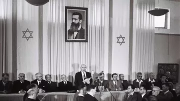 L'image montre une scène à l'intérieur d'une salle de réunion. Au fond, il y a un grand portrait en noir et blanc d'un homme, probablement une figure historique importante, entouré d'étoiles de David. Sur la scène, plusieurs hommes sont assis, en costume, tandis qu'un orateur se tient debout, s'adressant à l'assemblée. L'ambiance semble formelle, indiquant un événement significatif. Les participants, qui sont assis face à la scène, apparaissent attentifs à la présentation en cours.