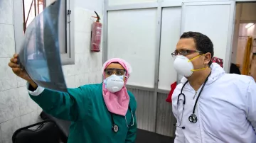 L'image montre deux professionnels de la santé examinant une radiographie. La femme porte un uniforme vert, un masque et un foulard rose, tandis que l'homme est vêtu d'une blouse blanche et porte également un masque. Ils se tiennent dans une pièce médicale, entourés de murs en carrelage. On peut voir un extincteur accroché au mur. L'expression de concentration indique qu'ils discutent sérieusement des résultats de l'examen.