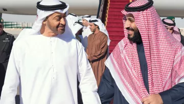 L'image montre deux hommes vêtus de vêtements traditionnels arabes, marchant côte à côte sur un tapis rouge. Ils semblent échanger un moment amical, souriant l'un à l'autre. En arrière-plan, plusieurs autres personnes sont présentes, ainsi qu'un avion stationné. L'atmosphère semble chaleureuse et détendue.