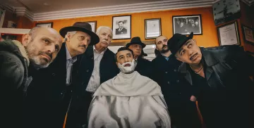 L'image montre un groupe de six hommes rassemblés autour d'un homme assis, qui est en train de se faire raser. L'homme au centre est allongé sur une chaise de barbier, avec de la mousse à raser sur le visage. Les membres du groupe sont vêtus de manteaux sombres et de chapeaux, affichant des expressions sérieuses et attentives. L'environnement semble être celui d'un barber shop classique, avec des cadres accrochés aux murs. L'atmosphère est à la fois mystérieuse et intrigante.