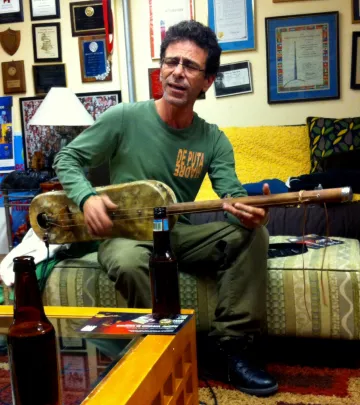 L'image montre un homme assis dans un intérieur chaleureux, probablement un salon. Il joue d'un instrument de musique, semblable à une guitare, tout en chantant. Il porte un t-shirt avec un message audacieux et des pantalons clairs. À côté de lui, sur une table basse, on aperçoit quelques bouteilles de bière. Les murs autour sont décorés de plaques et de diplômes, et un canapé coloré est visible en arrière-plan. L'ambiance semble conviviale et détendue.