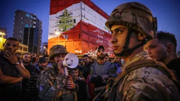 L'image montre un groupe de personnes rassemblées, dont plusieurs soldats en uniforme. L'un des soldats utilise un mégaphone, probablement pour communiquer avec la foule. En arrière-plan, on peut voir un bâtiment peint avec les couleurs du drapeau libanais. L'atmosphère semble tendue et engagée, suggérant une situation de manifestation ou de rassemblement important. La lumière est douce, indiquant peut-être le crépuscule. Les expressions des participants sont variées, allant de la concentration à l'écoute attentive.