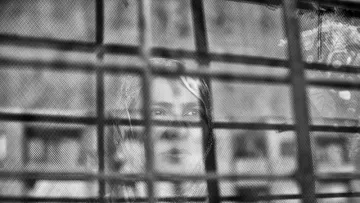 L'image montre une femme qui apparaît derrière une grille. Le contraste entre le flou et les barreaux de la grille donne une impression de séparation ou d'enfermement. La femme semble contemplative, et son expression peut évoquer des émotions profondes. Les nuances de noir et blanc ajoutent une atmosphère mélancolique à la scène.