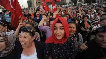 L'image montre une grande foule de personnes rassemblées, probablement lors d'une manifestation. Beaucoup d'entre elles tiennent des drapeaux turcs. On peut voir des expressions de passion et d'engagement sur les visages des participants. Certains semblent crier ou exprimer des émotions fortes. L'arrière-plan présente des bâtiments modernes, et il y a des affiches affichant des symboles ou des images en rapport avec la Turquie. L'atmosphère générale semble dynamique et pleine d'énergie collective.
