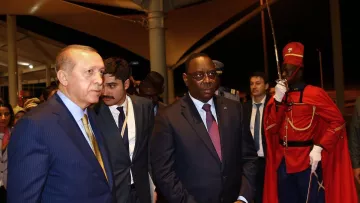 L'image montre deux hommes en costume, marchant ensemble dans un cadre officiel. L'un d'eux semble être un dirigeant turc, tandis que l'autre représente le Sénégal. Ils sont entourés de plusieurs personnes en arrière-plan, et un garde d'honneur en uniforme rouge se tient à proximité. L'atmosphère paraît sérieuse et formelle, typique d'une rencontre diplomatique.