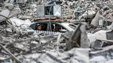 L'image montre un paysage dévasté avec des décombres sur le sol. Au centre, on aperçoit une voiture dans laquelle se trouvent deux personnes. La voiture semble être coincée parmi les débris, qui comprennent des morceaux de béton et d'autres matériaux d'une structure effondrée. L'ambiance générale dégage une impression de destruction et de chaos, témoignant d'une situation difficile.