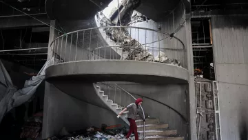 L'immagine mostra una scala a chiocciola danneggiata all'interno di un edificio in rovina. La struttura presenta macerie sparse e parte del soffitto è crollato, creando un'apertura sopra la scala. Una persona, vestita con un abbigliamento rosso e coperta da un copricapo, sta camminando sulla scala tra le macerie. L'atmosfera è quella di un luogo in crisi, con segni evidenti di distruzione e abbandono.