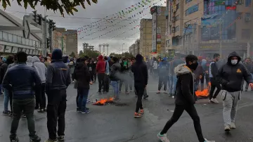 تظهر الصورة مجموعة كبيرة من الناس في منطقة حضرية، حيث يبدو أن هناك تظاهرة أو تجمع حاشد. يتواجد المتظاهرون في الشارع، وبعضهم يرتدي أقنعة وجه. هناك دخان يتصاعد من نيران صغيرة مشتعلة على الأرض. السماء تبدو غائمة، وهناك زينة ملونة معلقة في الجو، مما يشير إلى وجود نشاطات أو احتفالات بجانب المظاهرات. الأجواء تعكس التوتر نتيجة الظاهر.