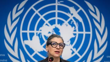 L'immagine mostra una donna che sta parlando di fronte a un microfono, con uno sfondo che presenta il logo delle Nazioni Unite. La donna ha gli occhiali e un'espressione seria, suggerendo che si tratta di una conferenza o di una presentazione ufficiale. Lo sfondo blu è decorato con il simbolo del globo terrestre, circondato da rami di ulivo, che rappresenta la pace.