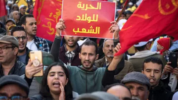 L'image montre une foule de manifestants, brandissant des drapeaux rouges. Au centre, un homme tient une pancarte avec le texte en arabe, tandis que d'autres personnes autour de lui semblent engagées dans la manifestation. On peut ressentir une atmosphère de protestation avec des expressions de détermination sur les visages des participants.