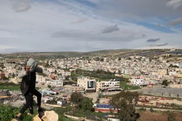 Nell'immagine si vede un giovane che si sta arrampicando su una roccia, con una città sullo sfondo. La città è composta da edifici di diversi piani, molti dei quali di colore chiaro. L'area è circondata da colline verdi e il cielo è parzialmente nuvoloso, suggerendo un clima variabile. Si possono notare anche alcune strutture agricole e aree di terreno aperto in primo piano.
