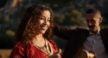 L'image montre une femme souriante avec des cheveux bouclés, portant une robe rouge ornée de motifs. Elle semble joyeuse et est entourée d'un environnement naturel. Un homme en arrière-plan, vêtu d'un costume, semble également profiter du moment. L'atmosphère générale est festive et conviviale.