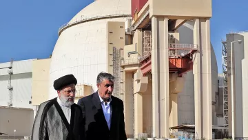 L'image montre deux hommes marchant devant une grande structure industrielle, qui semble être un réacteur nucléaire. Le bâtiment a une forme arrondie et est de couleur claire, avec des éléments de construction en béton visibles. Les deux hommes portent des vêtements formels ; l'un est vêtu d'un costume traditionnel et l'autre porte une veste. L'arrière-plan est dégagé, avec un ciel bleu. Cette scène pourrait illustrer une visite officielle ou une discussion liée à l'énergie nucléaire.