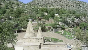 L'image montre un paysage montagneux avec une structure architecturale distinctive au centre. Cette structure présente deux tours ou pinacles coniques, typiques d'un style architectural particulier. Autour, on aperçoit des bâtiments en pierre et en béton, ainsi que des arbres verdoyants qui ajoutent une touche de nature. Le terrain est légèrement accidenté, avec des collines à l'arrière-plan, créant un environnement pittoresque et tranquille.