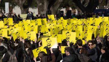 L'image montre une grande foule rassemblée, tenant des pancartes jaunes avec un visage représenté en noir. Les participants semblent mobilisés pour une cause, se tenant la main levée en signe de protestation ou de soutien. En arrière-plan, on peut voir d'autres personnes et des arbres, suggérant que l'événement a lieu en plein air. L'atmosphère paraît engagée et déterminée.