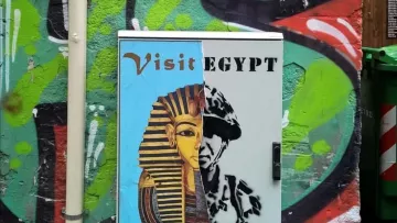 L'immagine mostra un armadietto metallico dipinto con un murale che presenta due volti distinti. A sinistra, c'è un'immagine stilizzata di un faraone, accompagnata da un invito a "Visit Egypt" e "Enjoy the". A destra, c'è un ritratto che sembra rappresentare un soldato o una figura autoritaria, con la parola "REGIME" sopra di esso. Lo sfondo è colorato e impreziosito da graffiti, creando un contrasto tra i due soggetti. La composizione sembra riflettere una critica sociale o politica legata all'Egitto.