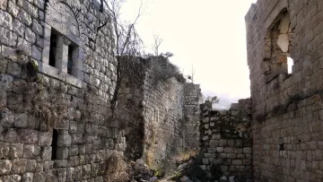 این تصویر نمایی از یک منطقه ویران‌شده است که دیوارهای سنگی و قدیمی آن هنوز پابرجا مانده‌اند. در اطراف دیوارها، گیاهان خشک و علف‌های هرز به چشم می‌خورند. روشنایی تصویر ممکن است نشان‌دهنده‌ی یک روز آفتابی باشد، با نور ملایمی که بر روی خرابه‌ها می‌تابد. برخی از دیوارها دارای پنجره‌های باز هستند و بخش‌هایی از ساختمان‌ها کاملاً تخریب شده‌اند. به طور کلی، جو تصویر حس خاطره‌ای از تاریخ و گذشته را منتقل می‌کند.
