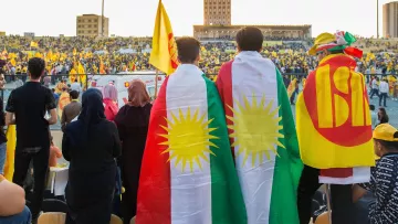 L'image montre un événement festif dans un stade rempli de supporters. Au premier plan, deux personnes portent des drapeaux, l'un étant le drapeau kurde avec les étoiles et l'autre avec un symbole qui semble représenter un mouvement ou un parti. Le public derrière eux est également vêtu de couleurs vives, principalement du jaune, suggérant une ambiance célébratoire. La scène évoque un fort sentiment d'unité et de fierté culturelle.