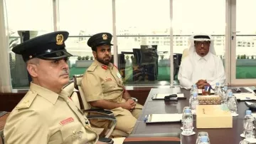 L'image montre un groupe de personnes en réunion dans un bureau moderne avec de grandes fenêtres offrant une vue sur l'extérieur. Il y a deux hommes en uniforme de police, portant des chapeaux et des uniformes beiges, qui semblent être en discussion. Un autre homme, vêtu d'une tenue blanche traditionnelle, est assis en face d'eux, semblant écouter attentivement. Des bouteilles d'eau sont présentes sur la table. L'atmosphère semble formelle et professionnelle.