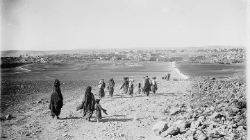 L'image montre un groupe de personnes marchant sur un chemin rural, entouré de paysages arides et de collines. Les individus, habillés de vêtements traditionnels, avancent ensemble, certains portant des charges sur la tête. En arrière-plan, on peut voir une ville avec des bâtiments, peut-être une petite agglomération, et une route qui serpente à travers le paysage. L'atmosphère semble sereine et la scène évoque un mode de vie traditionnel.