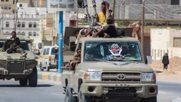 L'image montre un véhicule tout-terrain de type pick-up, spécifique à des zones de conflit, avançant sur une route. À l'arrière du camion, plusieurs personnes sont assises, certaines tenant des armes, suggérant une situation de tension ou de guerre. En arrière-plan, on peut apercevoir des bâtiments et des infrastructures qui semblent typiques d'une zone urbaine affectée par des conflits. La scène évoque un environnement militaire ou paramilitaire.
