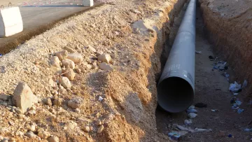 تظهر الصورة مشهدًا لموقع عمل حيث تم حفر خندق في أرض جافة. في وسط الخندق، يوجد أنبوب كبير على الأرض، ويبدو أنه يتم إعداده للتركيب. يحيط بالمنطقة بعض الحصى والصخور، مما يدل على أعمال البناء. في الخلفية، يمكن رؤية معدات بناء تظهر أنها تعمل في الموقع. كما يوجد بعض المخلفات والقمامة حول الخندق، مما يشير إلى تواجد نشاط بشري.