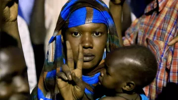 L'image montre une femme portant un foulard coloré, qui fait un signe de paix avec sa main. Elle tient un jeune enfant dans ses bras. L'arrière-plan semble flou, suggérant qu'elle se trouve dans une foule. Les expressions sur son visage et sa posture peuvent indiquer un moment de détermination ou de solidarité.