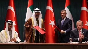 L'immagine mostra una cerimonia ufficiale con quattro uomini in abiti formali. Sullo sfondo si vedono le bandiere degli Emirati Arabi Uniti e della Turchia. Due degli uomini sono in piedi e sembrano scambiarsi documenti, mentre gli altri due sono seduti. L'atmosfera suggerisce un momento importante, probabilmente relativo a un accordo o a una collaborazione tra i due paesi.