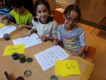 L'image montre deux jeunes filles assises à une table, souriant et travaillant sur des feuilles de papier. Elles utilisent des pinceaux et de l'encre, et semblent engagées dans une activité créative, probablement de calligraphie. Des étiquettes jaunes sont visibles, sur l'une est écrit "سيماء" et sur l'autre "دنيا" en arabe. L'environnement est scolaire, avec d'autres enfants visibles en arrière-plan.